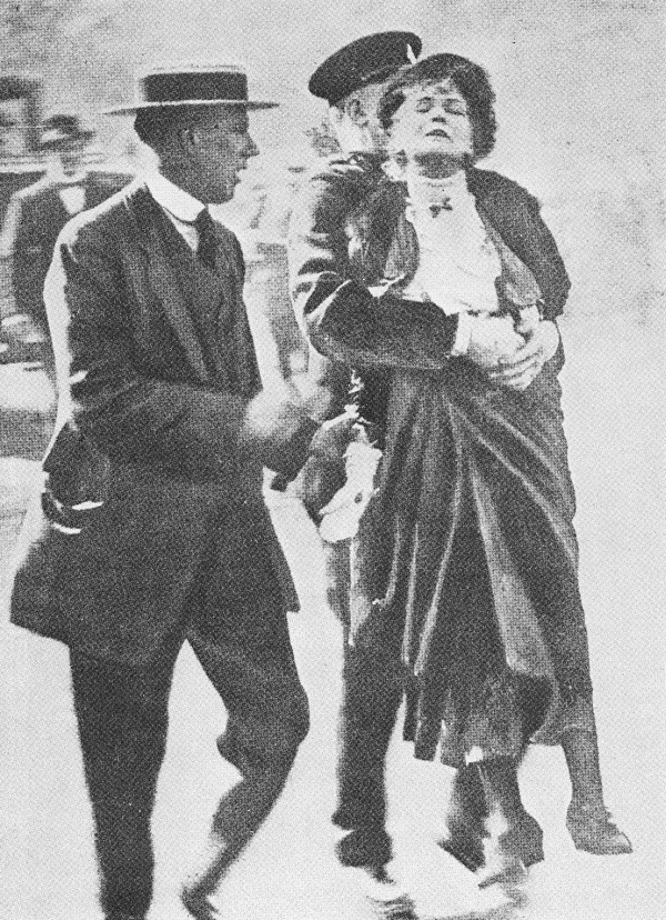 Mehrere Polizisten tragen die geschwächte Emmeline Pankhurst gewaltsam durch eine dichte Menschenmenge.